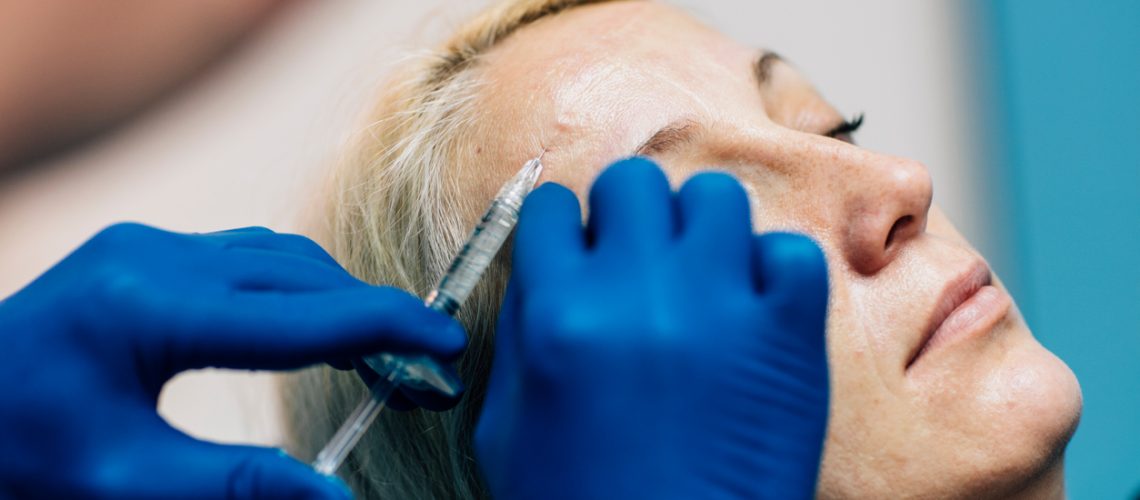 Doctor wears blue surgery gloves injecting hyaluronic acid under patients skin on forehead to fix facial wrinkles. Face Lifting Treatment. Patient is mature adult woman in her early 40s. Selective focus to syringe going under the skin. Real process has been shot. Canon EOS 5Ds.